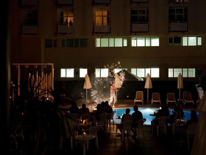 Nachtaufnahme eines beleuchteten Pools mit Gästen und Sonnenschirmen neben einem Hotelgebäude.