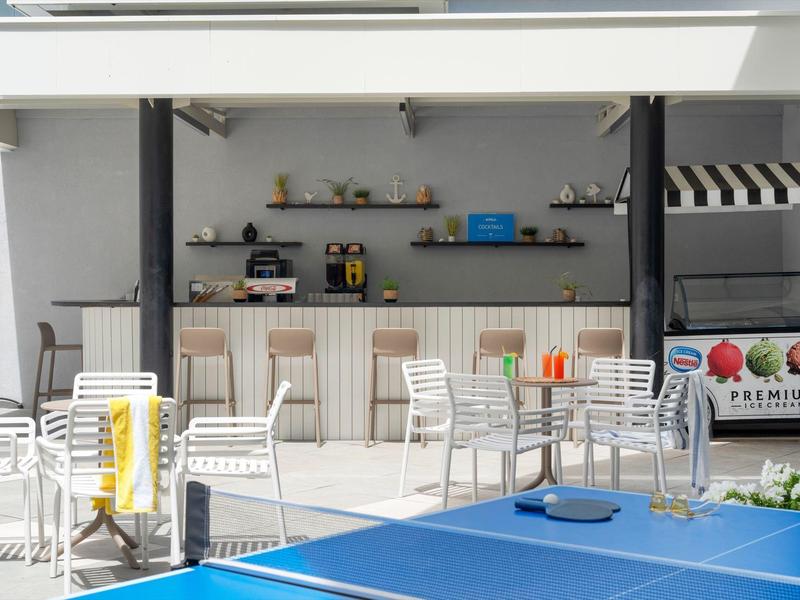 Freizeitbereich mit Tischtennisplatte, weißen Stühlen und Bar mit Getränken im Hintergrund.