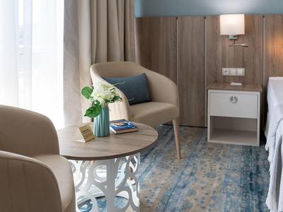 Gemütliche Sitzecke mit zwei beigen Sesseln, rundem Tisch, Vase und blau-grauem Teppich neben Bett.