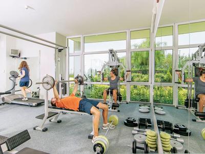 Sala fitness con tre persone che si allenano su macchine e grandi finestre che si affacciano sul verde.