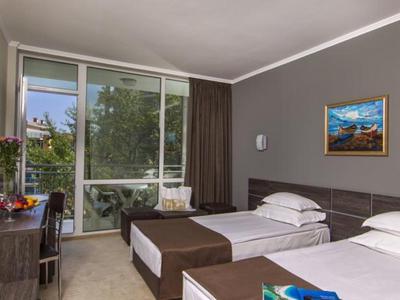 Hotelzimmer mit zwei Einzelbetten, Schreibtisch, Fernseher und großer Fensterfront mit Vorhängen.