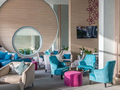 Moderne Hotellobby mit blauen Sesseln, rosa Hockern und rundem Spiegel an Holzpaneelwand.