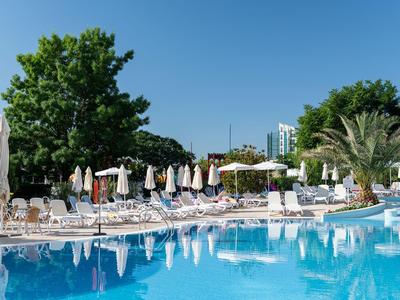 Großer Swimmingpool mit weißen Liegestühlen und Sonnenschirmen vor grünen Bäumen und blauem Himmel.
