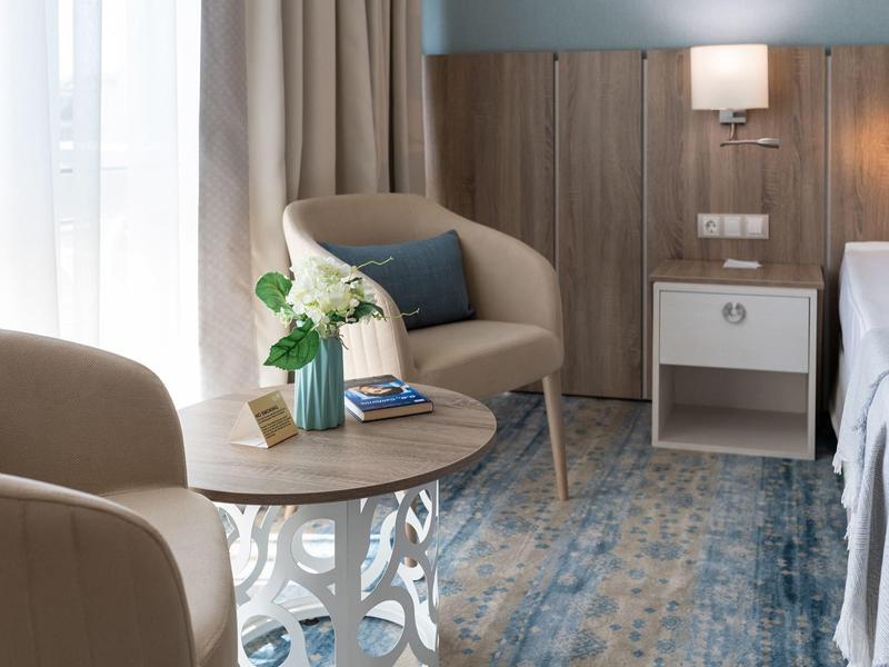 Gemütliche Sitzecke mit zwei beigen Sesseln, rundem Tisch, Vase und blau-grauem Teppich neben Bett.