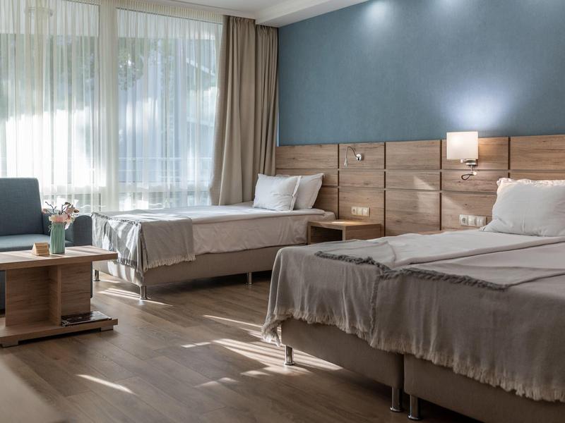 Hell eingerichtetes Zimmer mit zwei Betten, Holzmöbeln, blauer Wand und großen Fenstern.