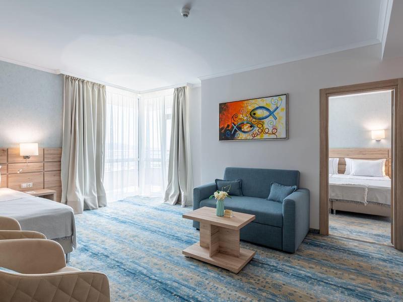 Helles Hotelzimmer mit Bett, Sofa, Teppich in Blau- und Grautönen, farbigem Bild und großem Fenster.