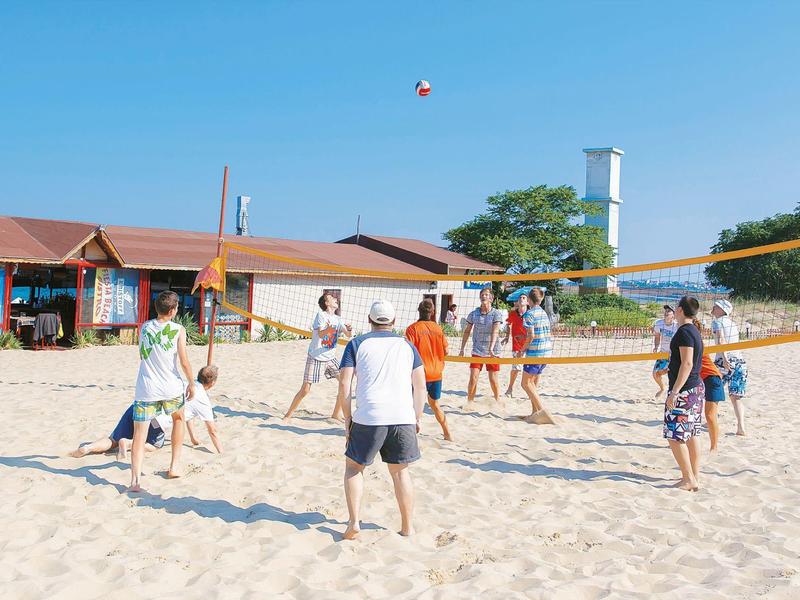 Menschen spielen Volleyball am Sandstrand vor einem Gebäude unter klarem blauem Himmel.