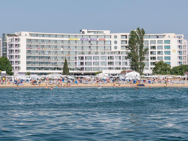 Großes weißes Hotelgebäude am Strand mit vielen Menschen und blauem Meer im Vordergrund.