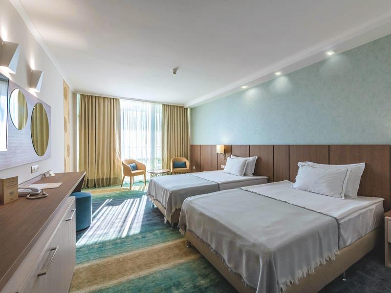 Helles Hotelzimmer mit zwei Betten, grünem Teppich, Holzpaneelen und großen Fenstern mit Vorhängen.