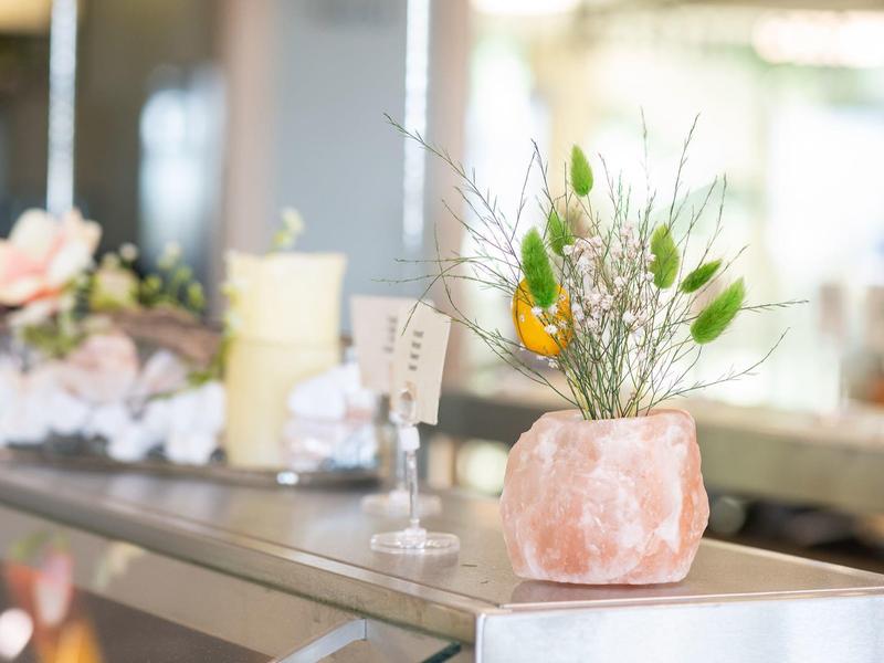 Grüne Zweige in rosa rosa Salzstein-Vase auf glänzendem Metalltisch in heller Raumumgebung.