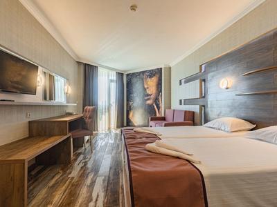 Modern eingerichtetes Hotelzimmer mit großem Bett, TV, Fenster mit Vorhängen und Holzfußboden.