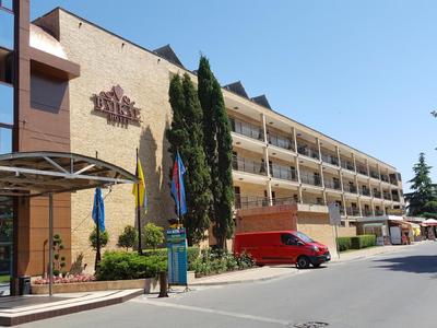 Mehrstöckiges Hotelgebäude mit Balkonen, Zypressen und einem roten Lieferwagen vor dem Eingang.