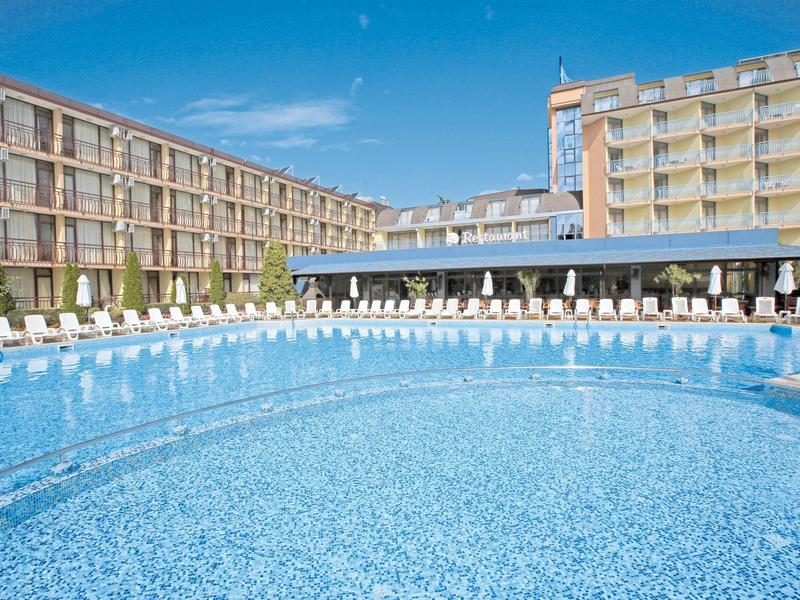 Groot hotelzwembad met ligstoelen en moderne gebouwen op de achtergrond onder een zonnige hemel.