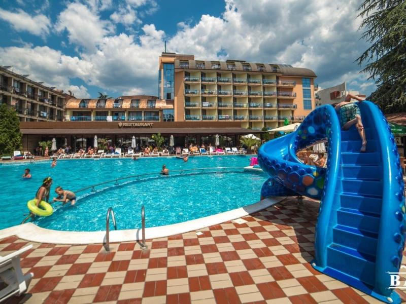 Hotelzwembad met blauwe glijbaan, gasten en meerlaagse hotel onder zonnige lucht.
