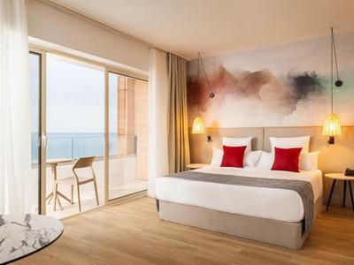 Habitación de hotel moderna con vista al mar, cama doble, pared artística y balcón con sillas.