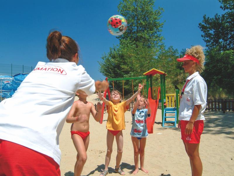 Niños y monitores juegan con una pelota en una zona arenosa con clima soleado.
