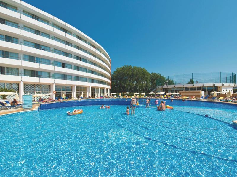 Gran piscina al aire libre frente a un edificio moderno de hotel con personas y sombrillas en un día soleado.