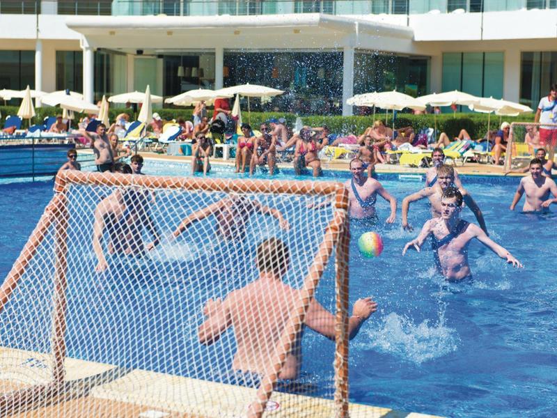 Personas jugando polo acuático en una piscina frente a un hotel con sombrillas blancas.
