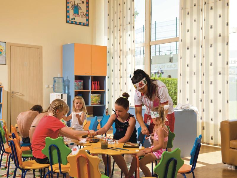 Niños sentados en mesas pequeñas coloreando en un aula luminosa con un supervisor.