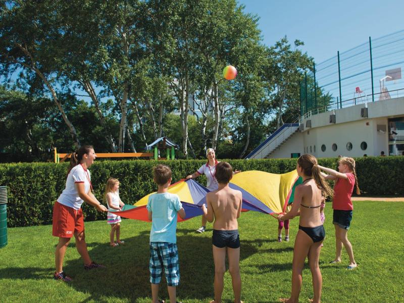 Niños juegan con un paracaídas colorido y una pelota en un césped verde con clima soleado.