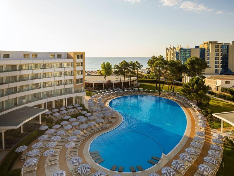 Hotel con piscina curvada y tumbonas junto a la costa al atardecer.
