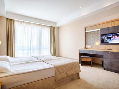 Lichte hotelkamer met tweepersoonsbed, bureau en aan de muur gemonteerde tv voor grote ramen met gordijnen.