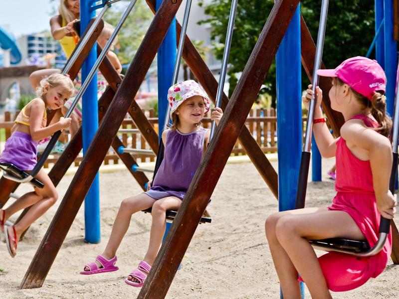 Drie kinderen schommelen op een zandige speelplaats op een zonnige dag.