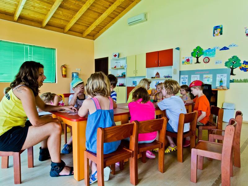 Kinderen zitten aan kleine tafels in een lichte, kleurrijke klas met een houten dak.