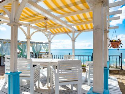 Terrasse mit weißem Holzmobiliar, gelb-weiß gestreiftem Sonnenschutz und Blick auf das blaue Meer.