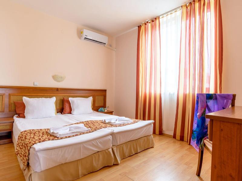 Bright hotel room with double bed, wooden floor, and curtains on the window.