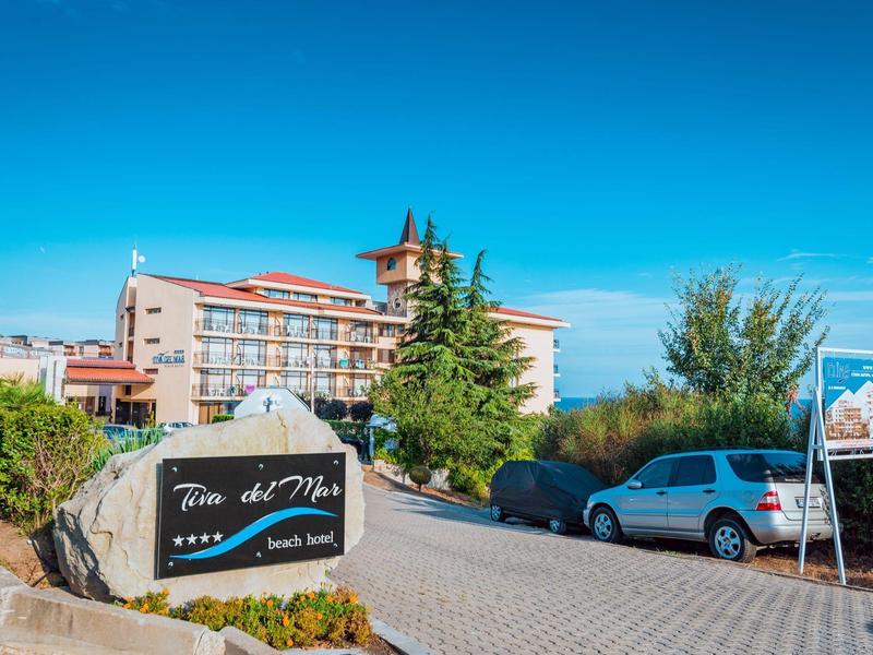 Hotel entrance with parking area, sign, and trees under a clear blue sky.