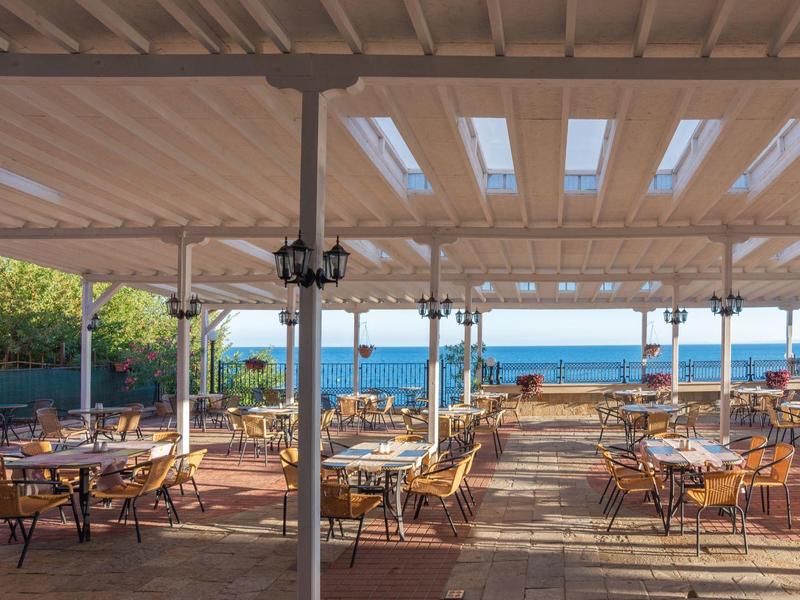 Covered terrace with tables and chairs overlooking the blue sea under a clear sky.