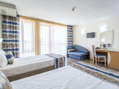 Hotel room with two single beds, curtained windows, desk, and sofa in a bright space.