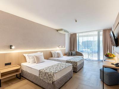 Jasny pokój hotelowy z dwoma podwójnymi łóżkami, biurkiem, telewizorem i balkonem z widokiem na taras.