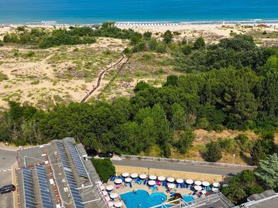 Widok na basen hotelowy z parasolami obok zalesionego terenu i wydm piaszczystych przy morzu.