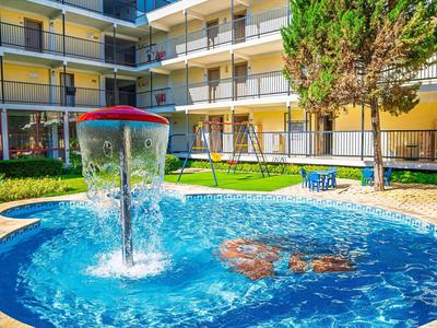 Mały okrągły basen z fontanną przed wielopiętrowym budynkiem hotelowym i terenem zielonym.