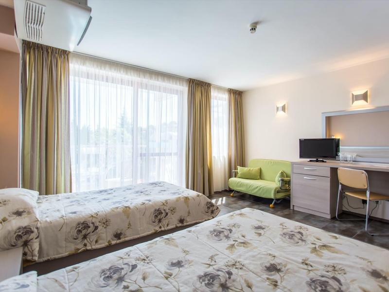 Hotel room with two beds, green sofa, desk, and large window with curtains.