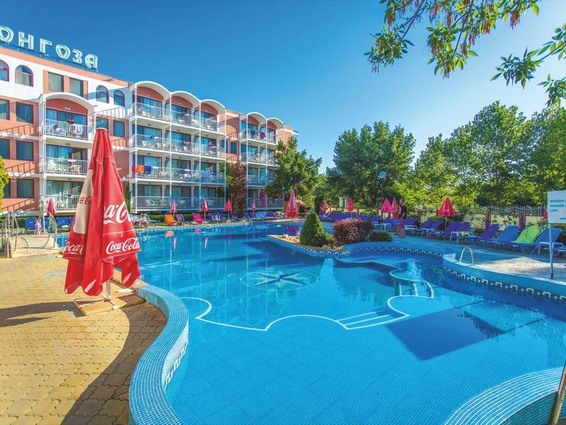Hotel building with balconies and swimming pool featuring red umbrellas and green trees under clear sky.