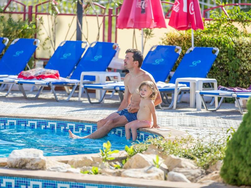 Man and child sitting at the edge of a pool with blue lounge chairs in the background.