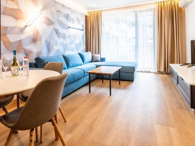 Helles Wohnzimmer mit blauem Ecksofa, Holzboden, großem Fenster und gedecktem Essplatz.