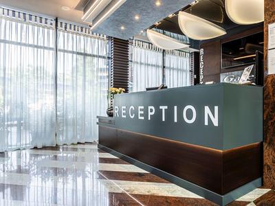 Moderne Hotellobby mit Rezeptionstresen, hellem Marmorfußboden und großen Fenstern mit Gardinen.