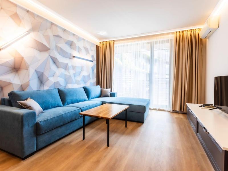 Helles Wohnzimmer mit blauer Eckcouch, Holzparkett, großem Fenster und moderner Tapete an der Wand.