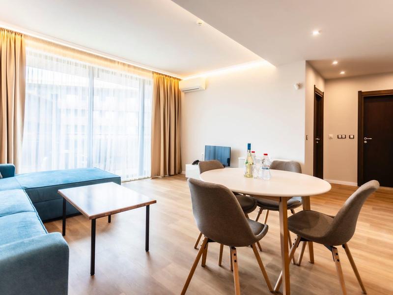 Helles Wohnzimmer mit blauen Sofas, rundem Holztisch, grauen Stühlen und großem Fenster mit Vorhängen.