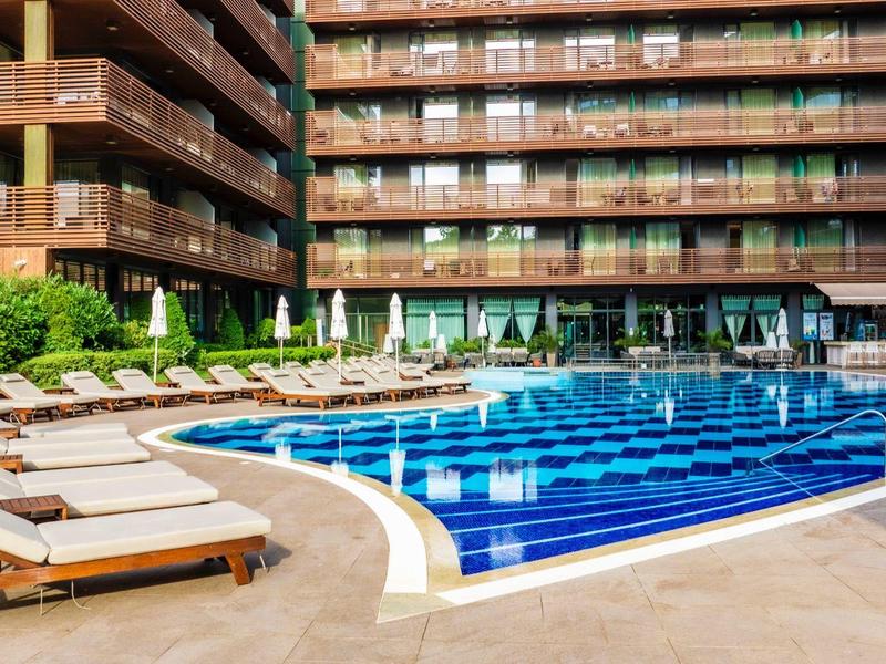 Geschwungener Außenpool mit Sonnenliegen vor einem mehrstöckigen Hotelgebäude.