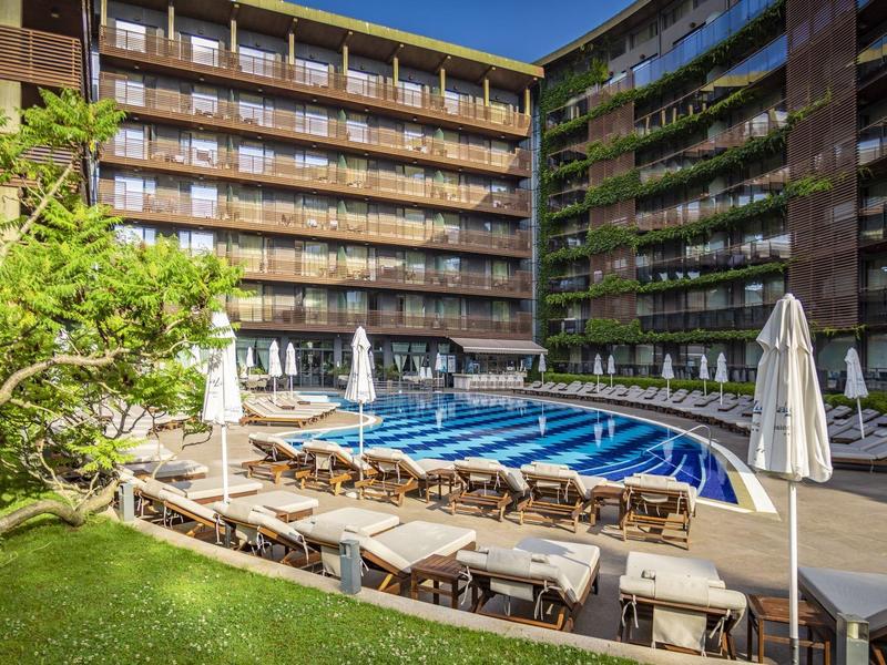 Hotel mit großem Außenpool, Liegestühlen und Grünflächen bei klarem Himmel.