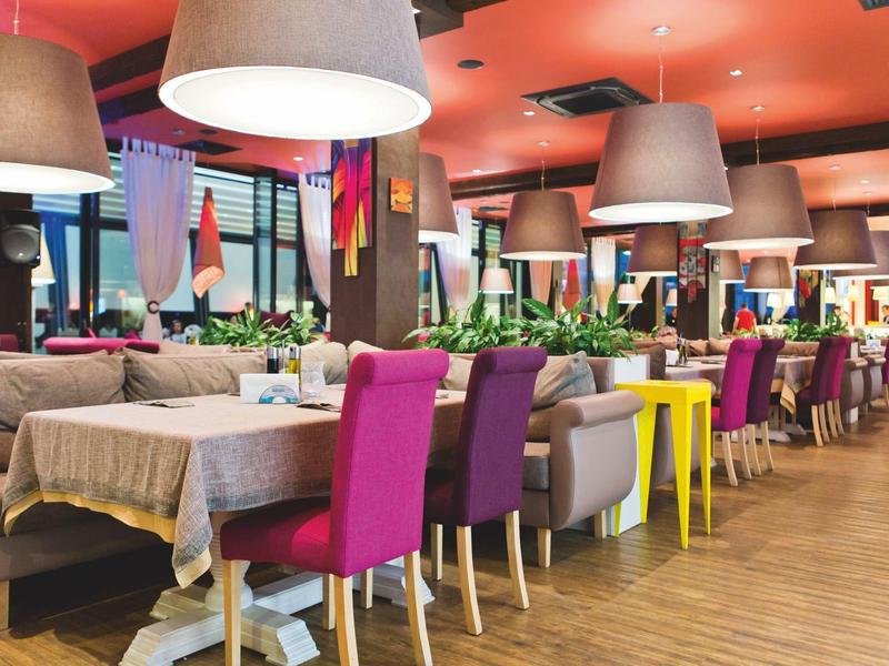 Moderner Gastraum mit bunten Stühlen, Tischdecken, großen Lampen und warmem Holzboden.