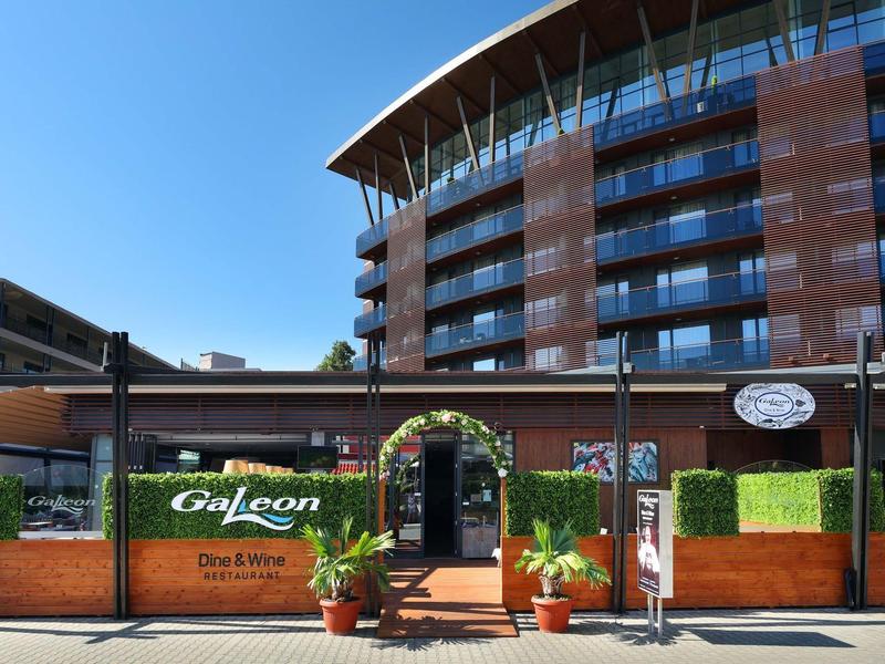 Modernes Hotel mit gebogenem Dach, großen Glasfenstern und einer Terrasse mit Pflanzen und Eingangsbogen.