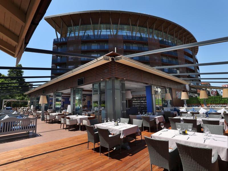 Außenbereich eines Restaurants mit gedeckten Tischen, Holzterrasse und rundem Glasgebäude im Hintergrund.