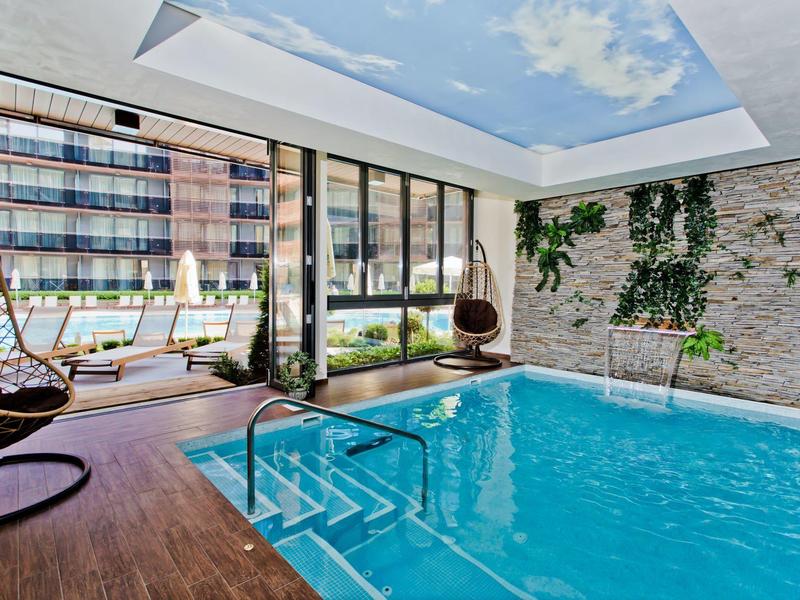 Moderner Innenpool mit Holzdeck, Glaswänden, grünen Pflanzen und Himmel-Deckenmalerei.