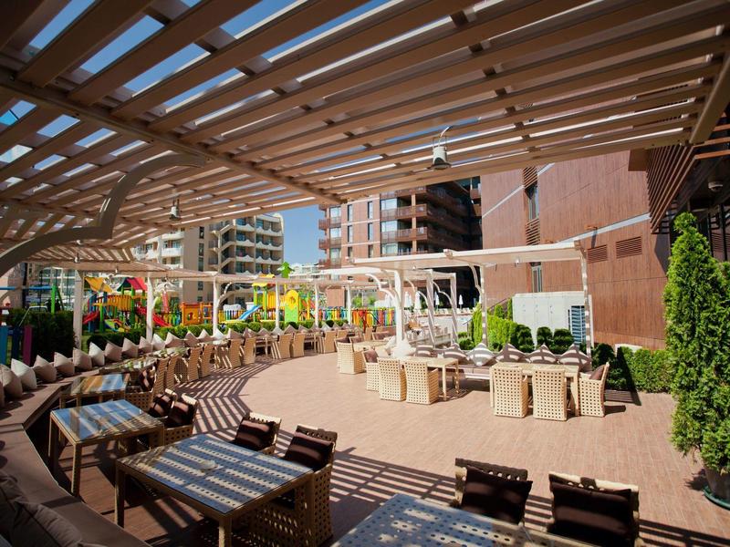 Große Terrasse mit Holzboden, Pergola, Sitzgruppen und grünen Pflanzen zwischen Wohngebäuden.